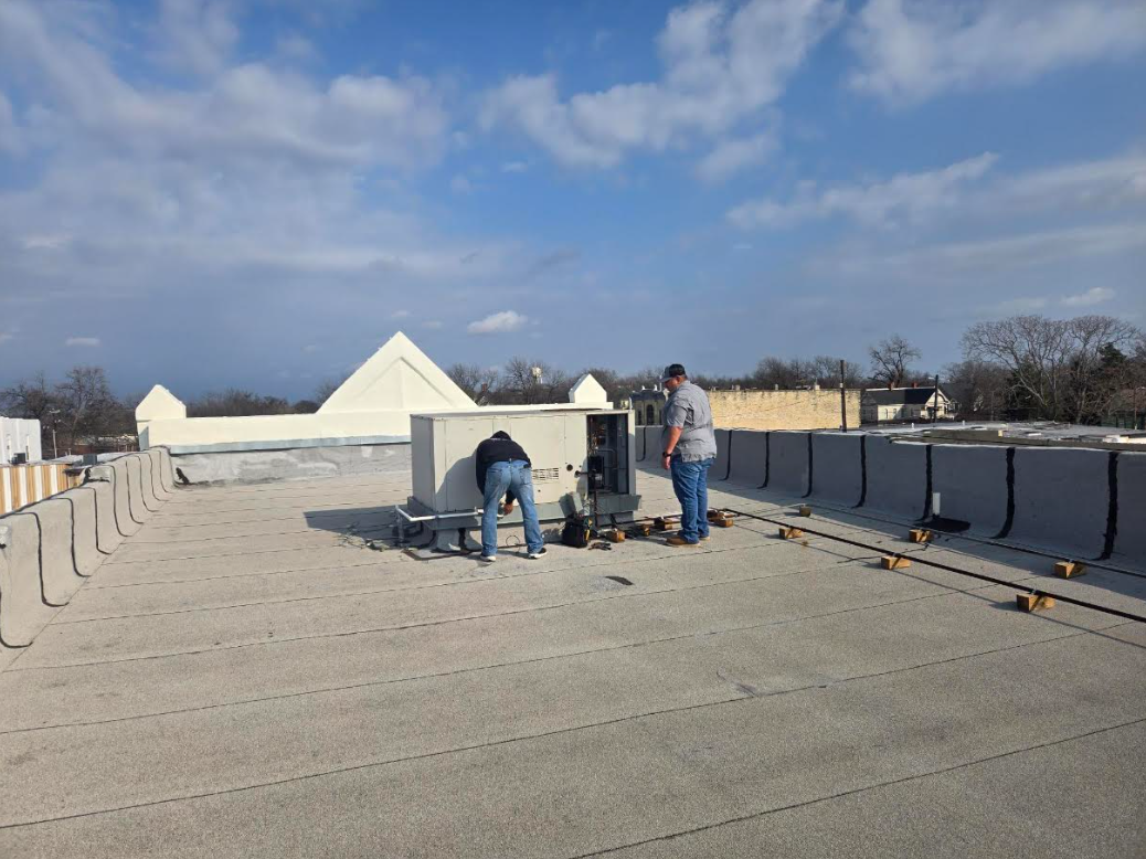 Workers on a flat rooftop installing materials beside white rooftop structures under a blue sky