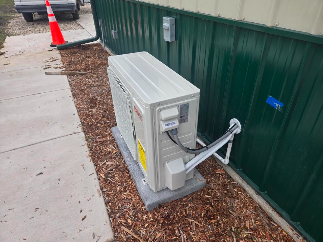Outdoor electrical unit beside a green fence, with conduit and a traffic cone nearby on a sidewalk.