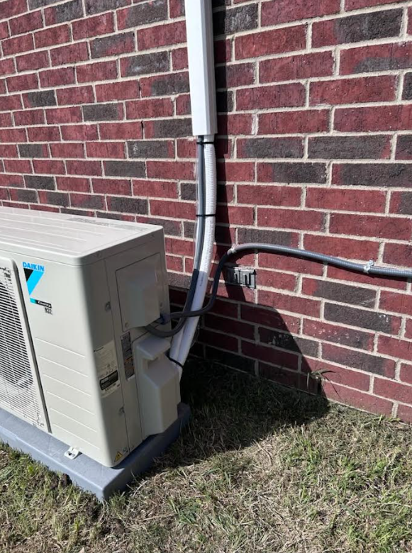 Outdoor HVAC unit beside a brick wall with a black pipe and conduit running upward.