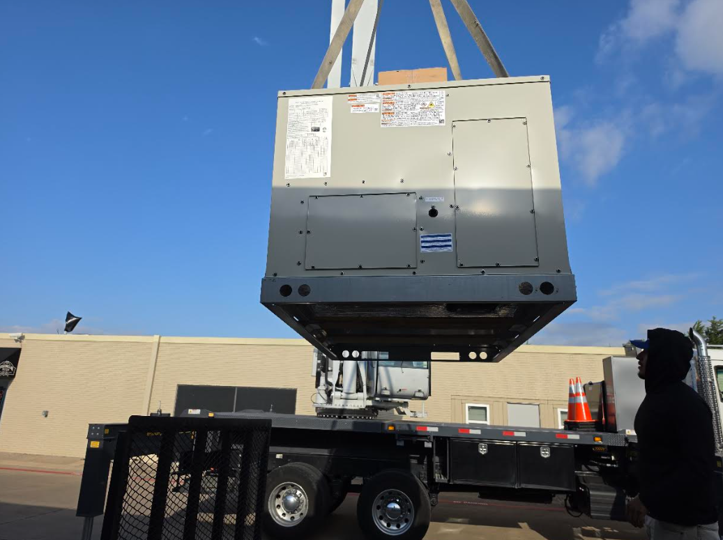 Large industrial machine suspended by crane above a flatbed truck in an outdoor lot