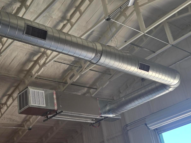 Industrial ceiling with silver ventilation ducts and a wall-mounted unit in a warehouse-like interior
