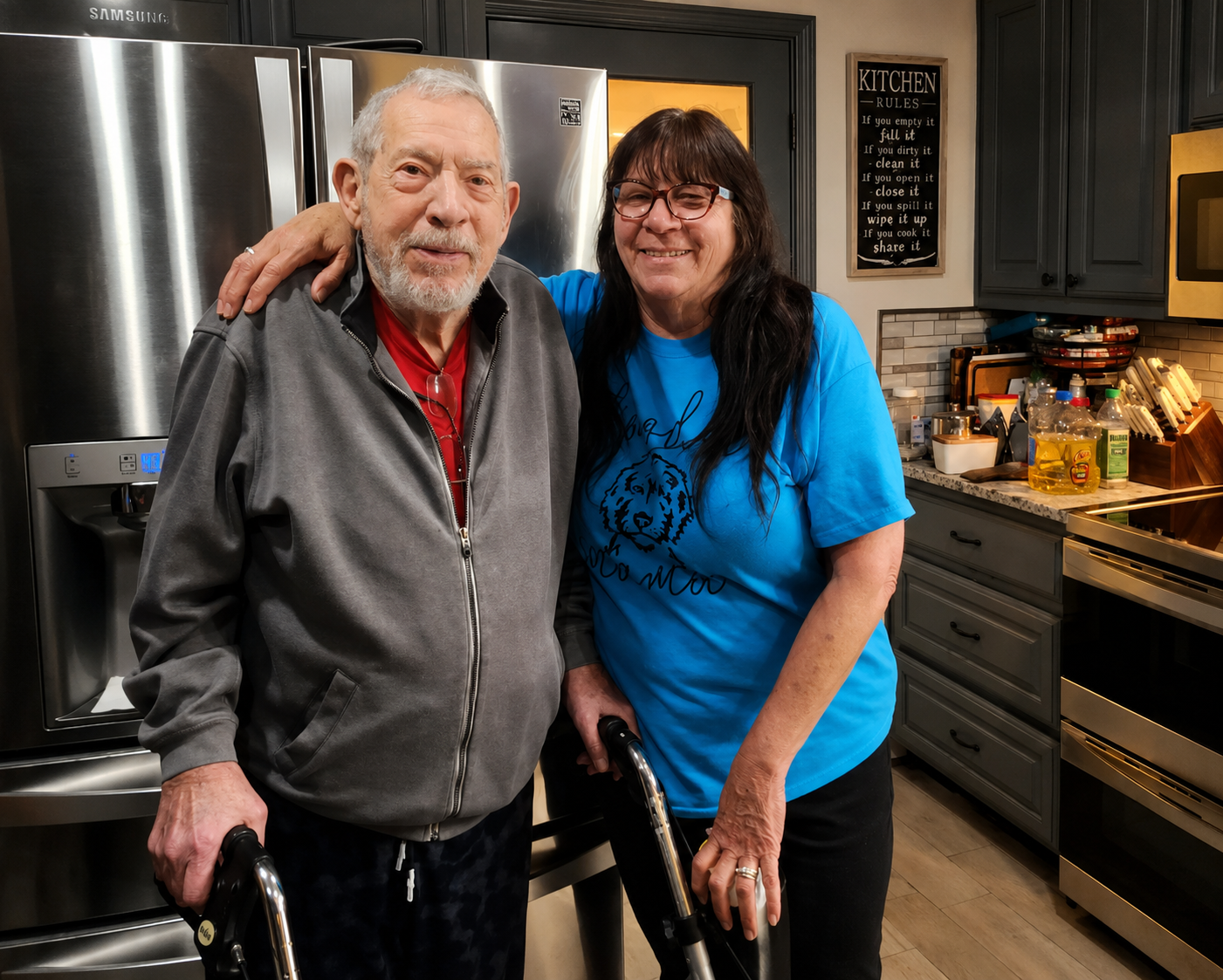 Two smiling people stand in a kitchen, one with a walker, beside stainless steel appliances.