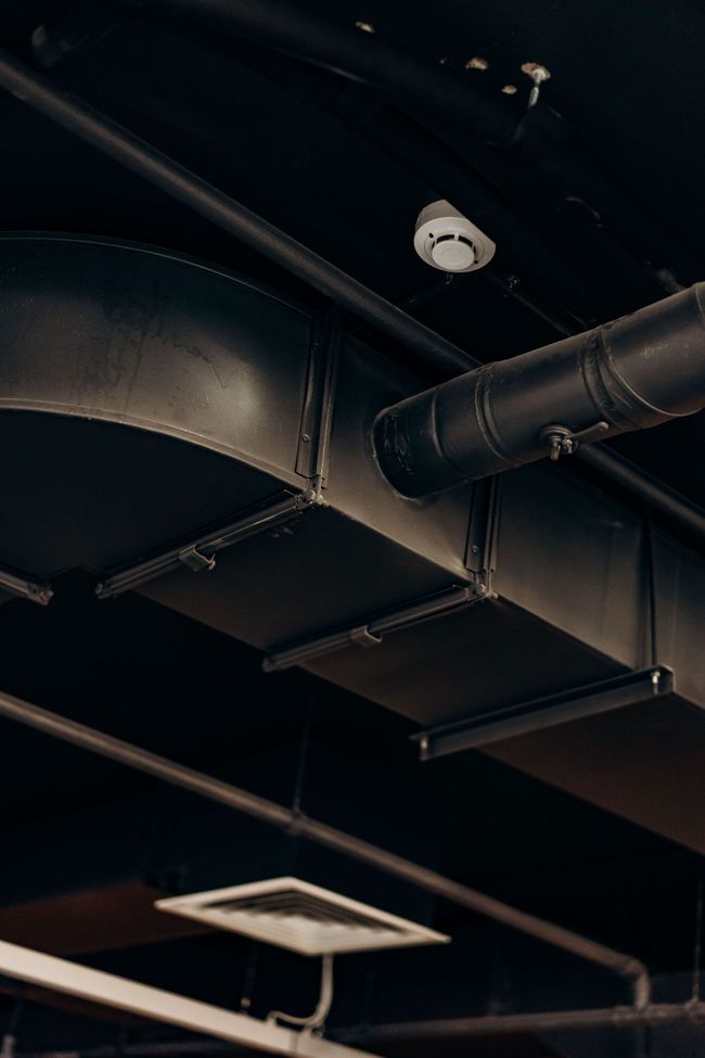 Ceiling with black pipes, ducts, and a small white fixture in an industrial interior
