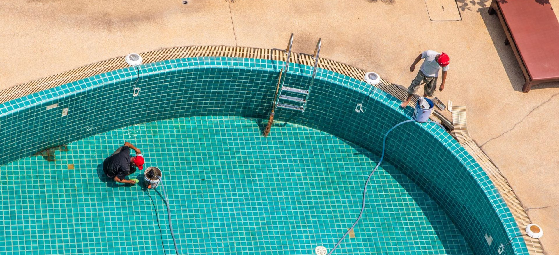 Workers cleaning a turquoise swimming pool with a hose and brush next to a tan deck