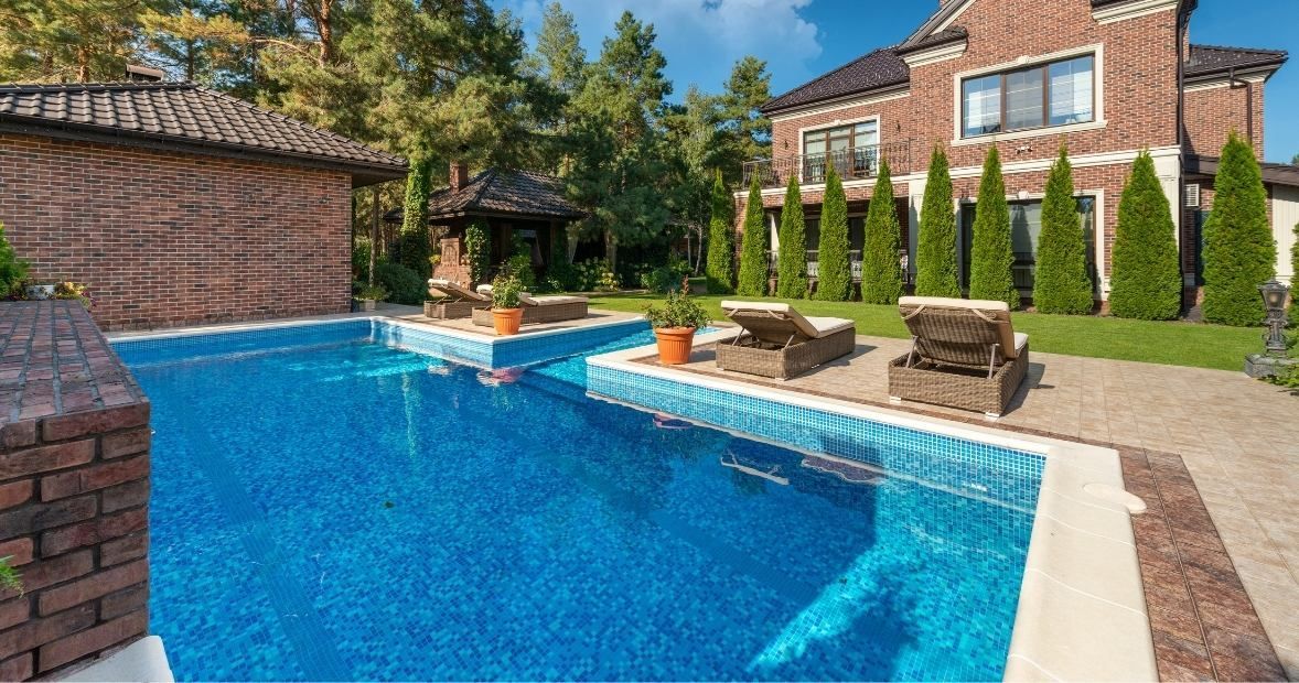 Backyard pool with blue water, brick patio, lounge chairs, and a brick house with tall evergreens