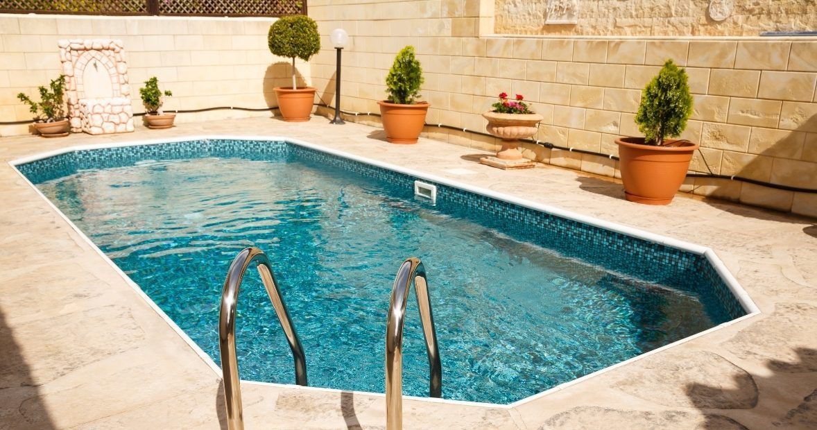 Small stone-tiled backyard pool with blue water, steps, and potted plants along the patio