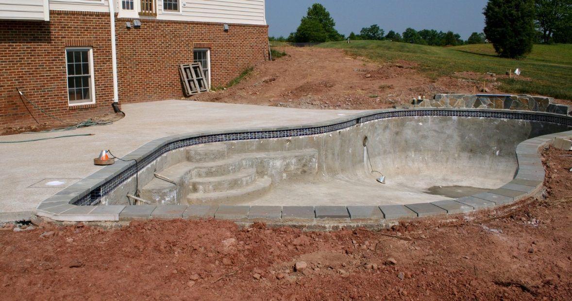 Empty in-ground swimming pool under construction beside a brick house, with dirt and concrete surrounding it