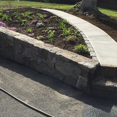 A stone wall with a concrete walkway leading to a flower bed.