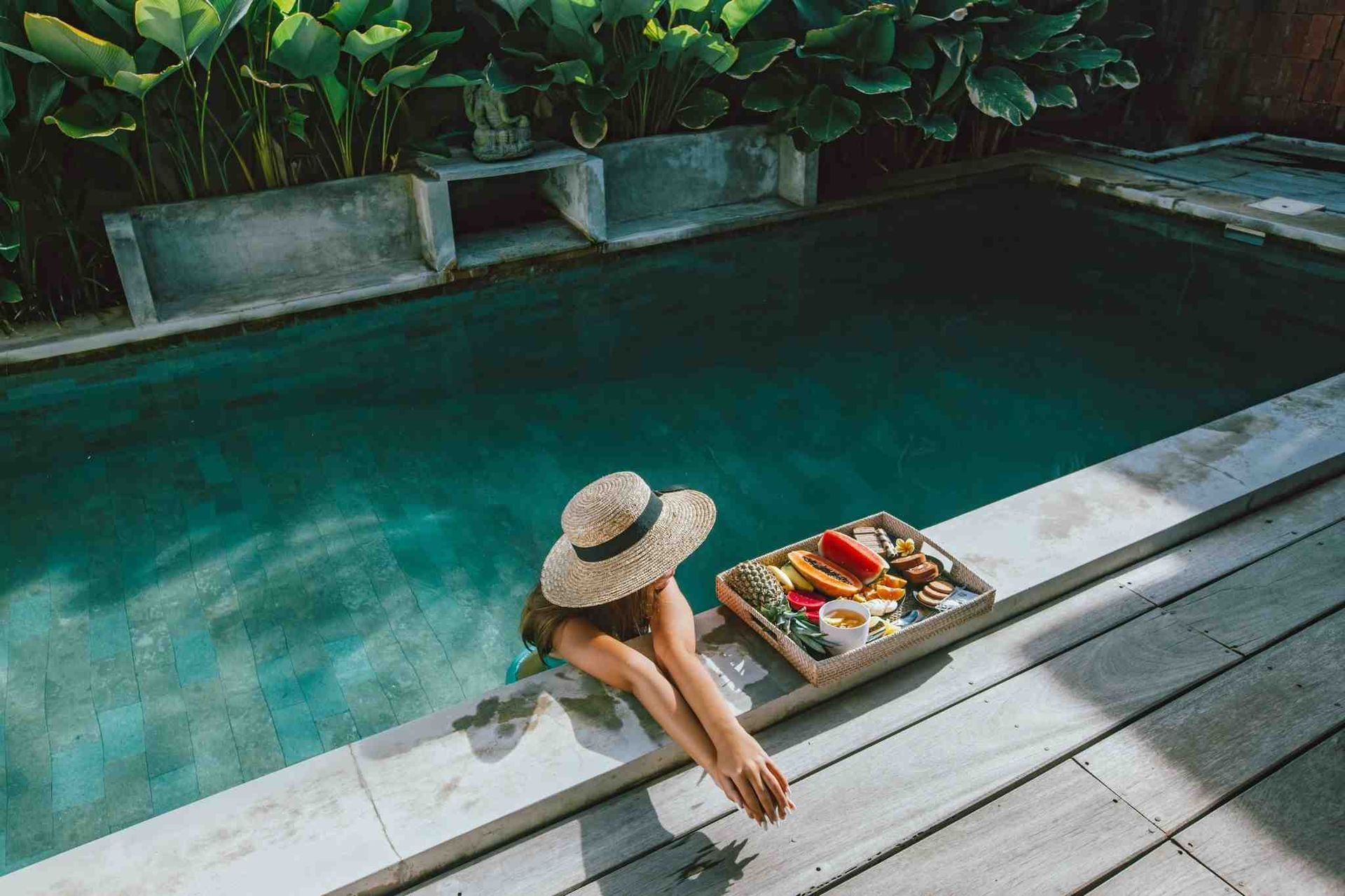 Woman in sun hat relaxing by a pool, enjoying fruit on a tray.