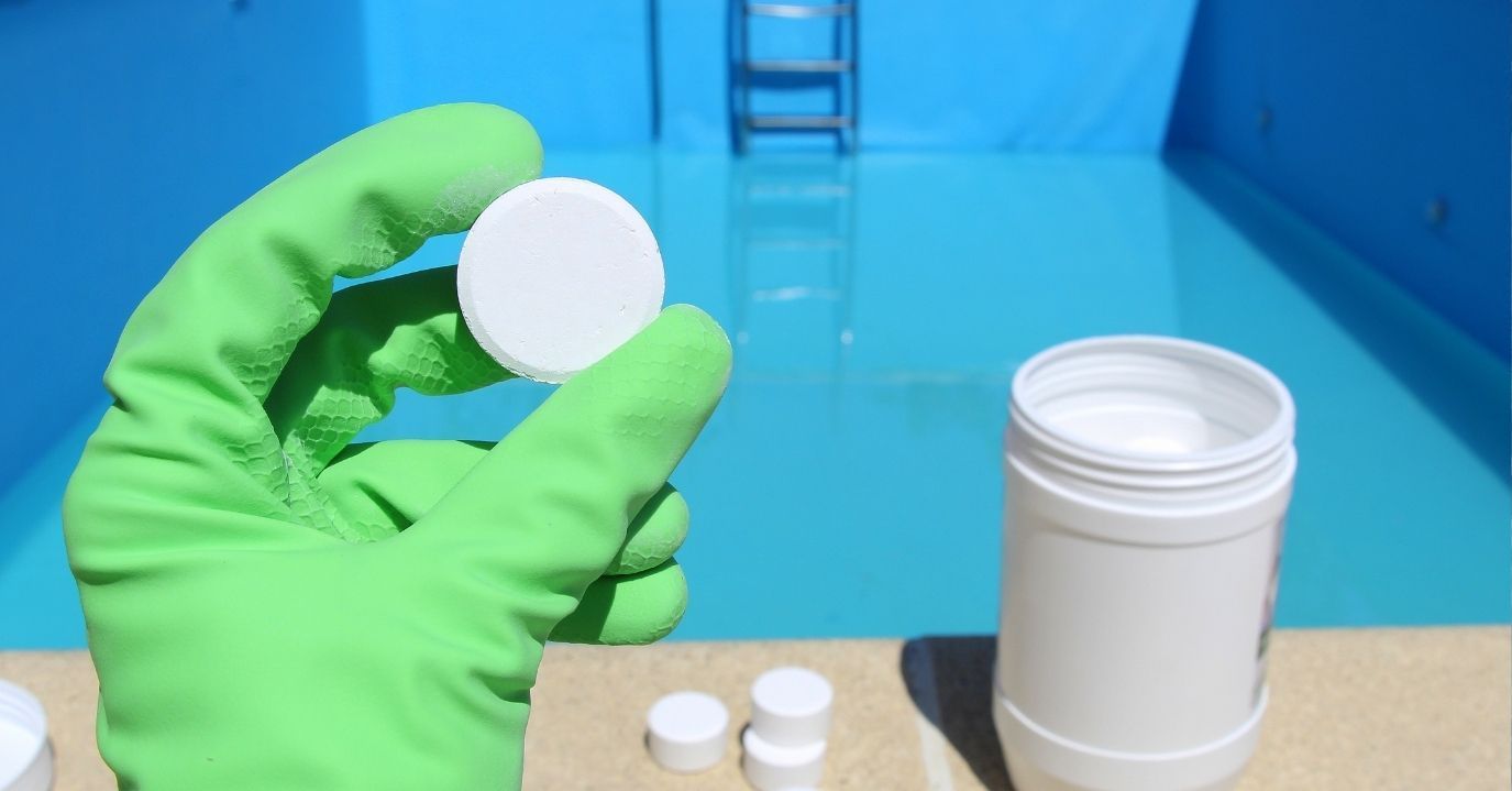 A hand in a bright green rubber glove holds a white, circular chlorine tablet