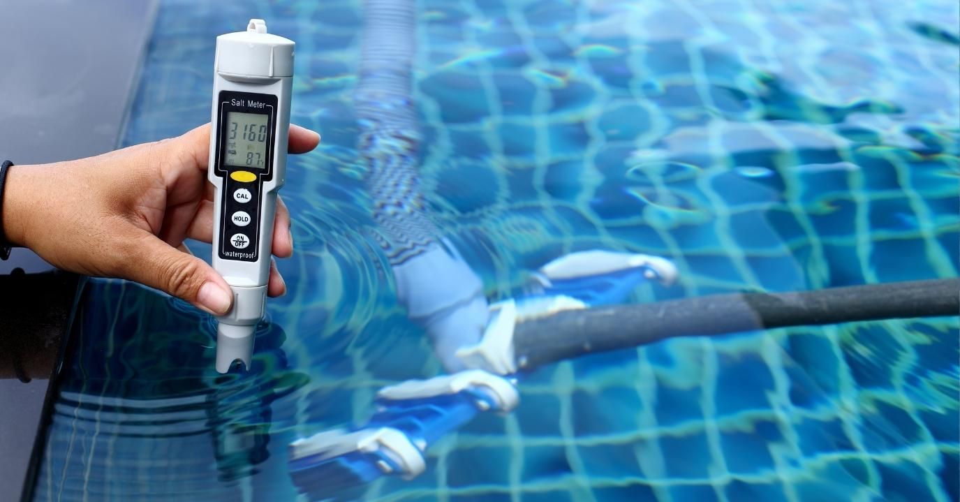 A hand holds a digital salinity meter over the edge of a swimming pool.