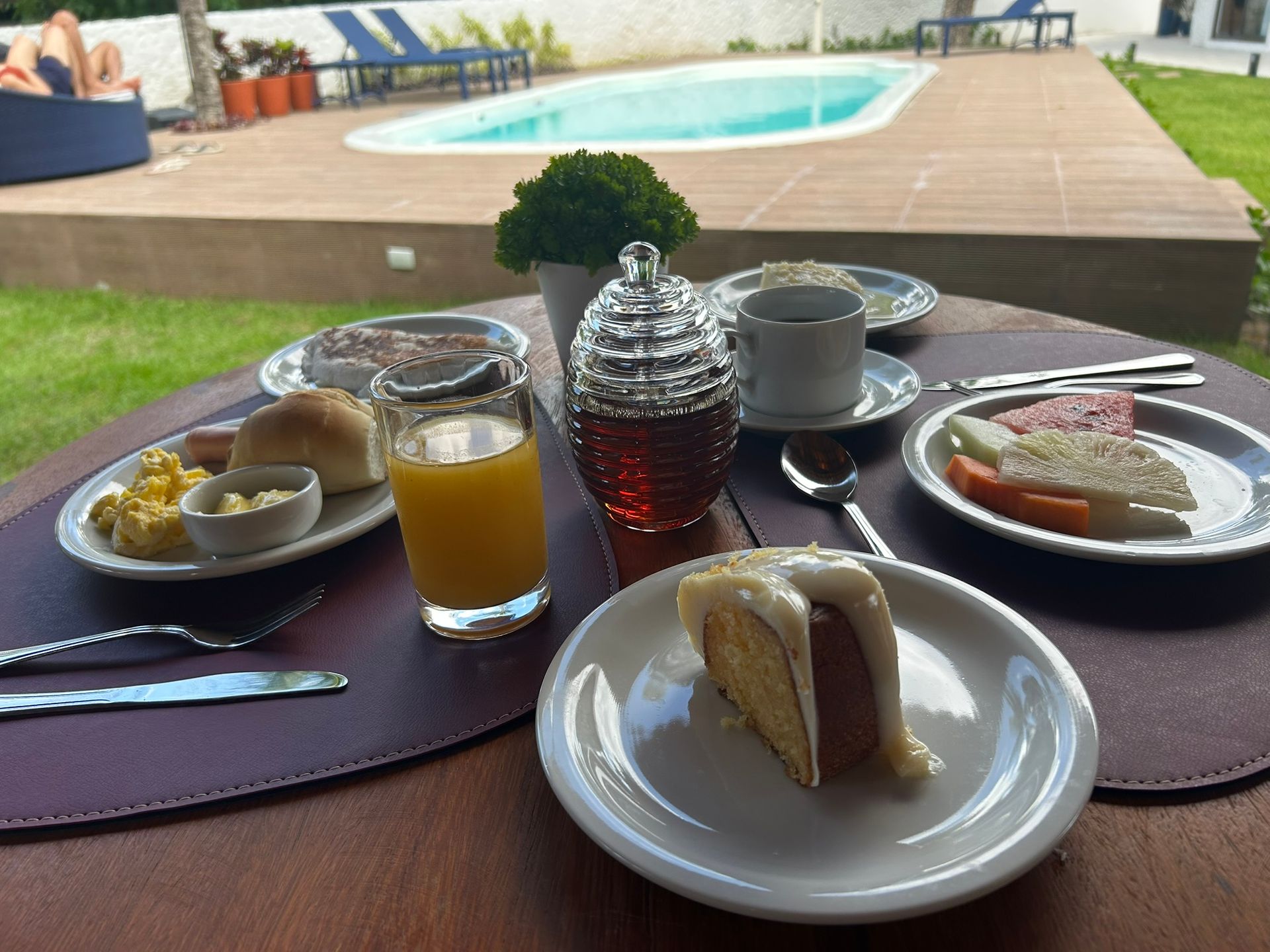 Uma mesa coberta com pratos de comida e bebidas com uma piscina ao fundo. Coralli Pousada