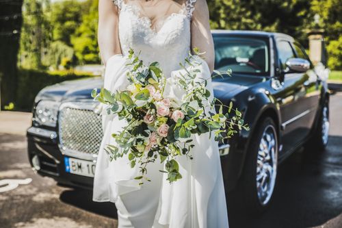 Bride holding white floral bouquet in front of a black luxury car outdoors