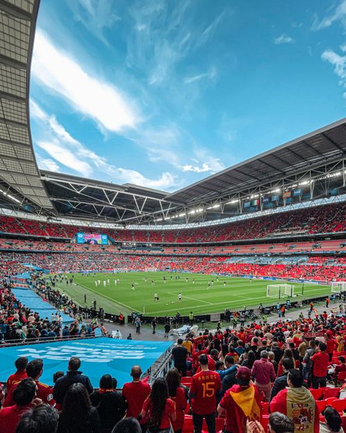 Crowded soccer stadium with a green pitch, red stands, and fans watching a match under a blue sky