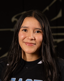Portrait of a smiling person with long dark hair wearing a black shirt, against a dark background.
