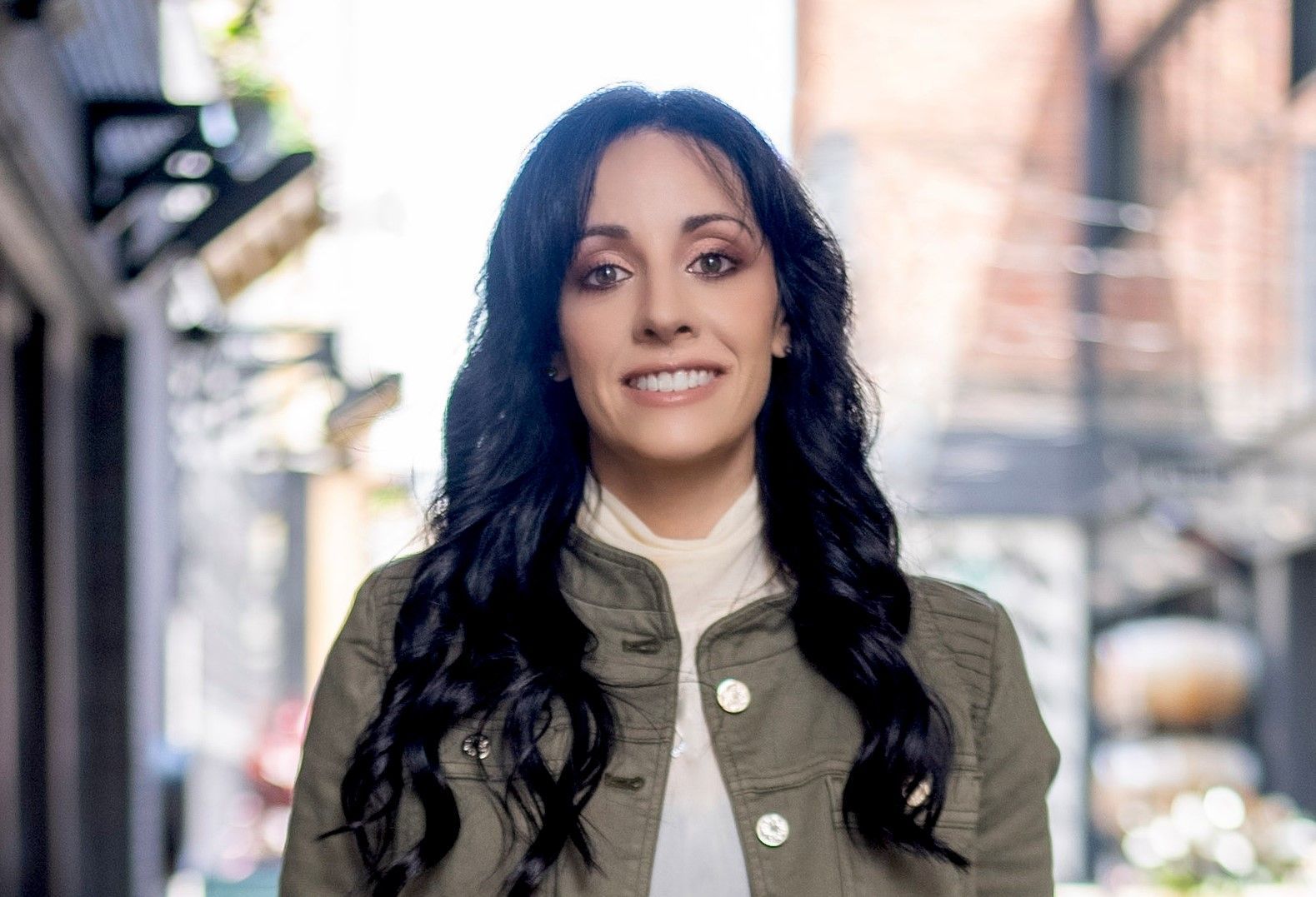 Smiling person with long dark hair in an olive jacket standing on a city street