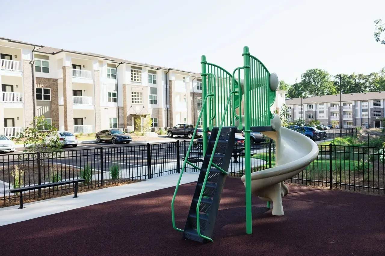 Green playground with a spiral slide in an apartment community courtyard, with a black fence and parked cars nearby.
