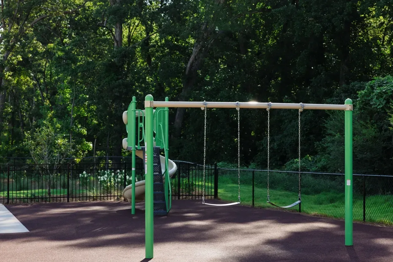 Green playground with swings and a slide, fenced area beside trees.
