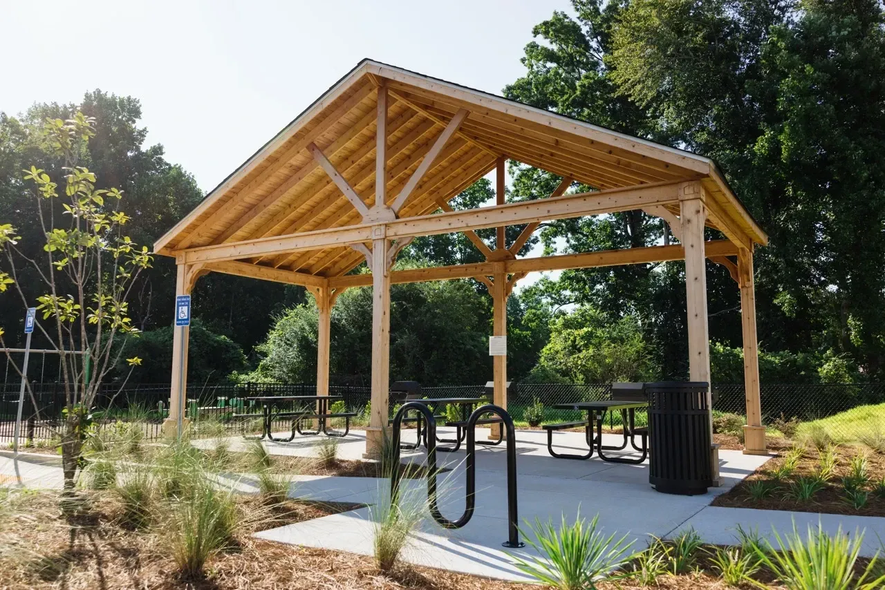 Open-air wooden pavilion with picnic tables in a landscaped outdoor area.