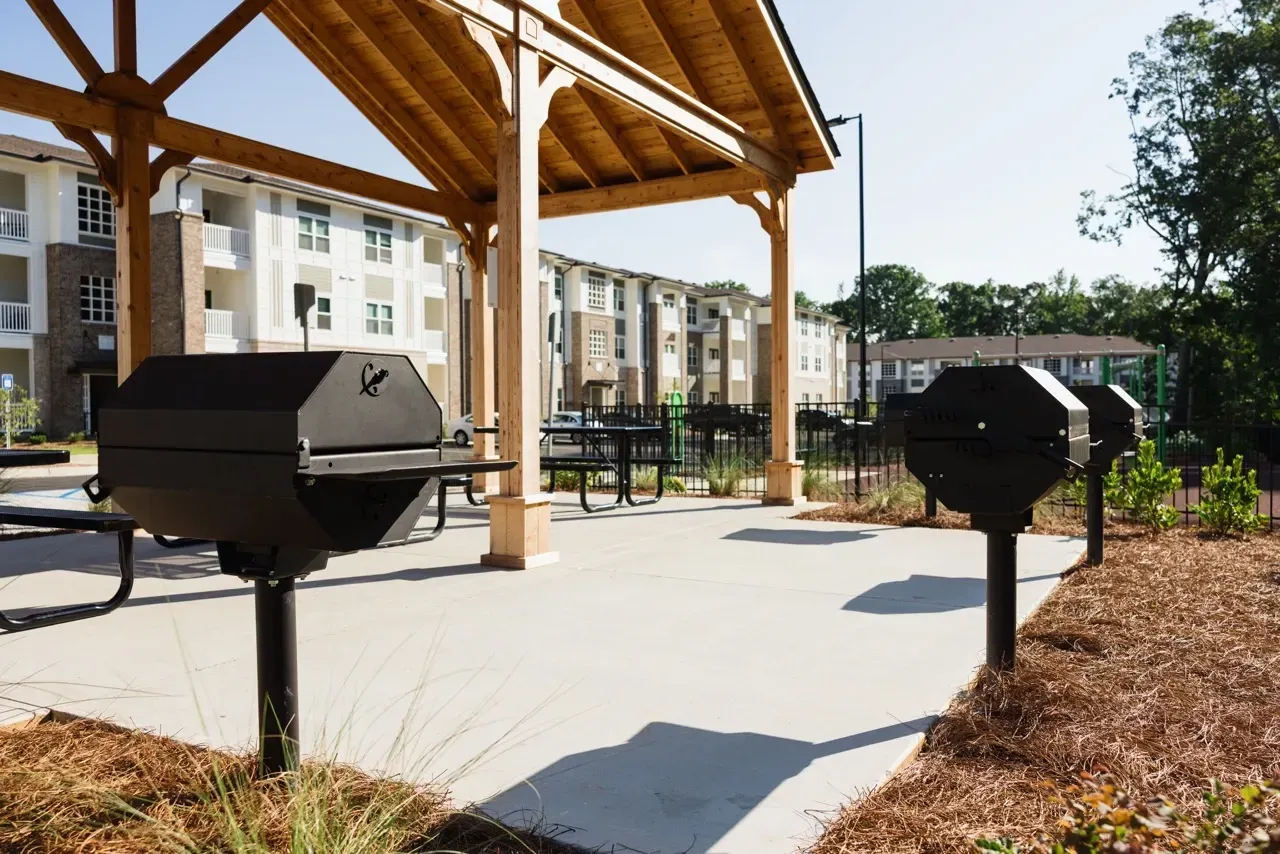 Outdoor community grilling area under a wooden pavilion beside apartment buildings.