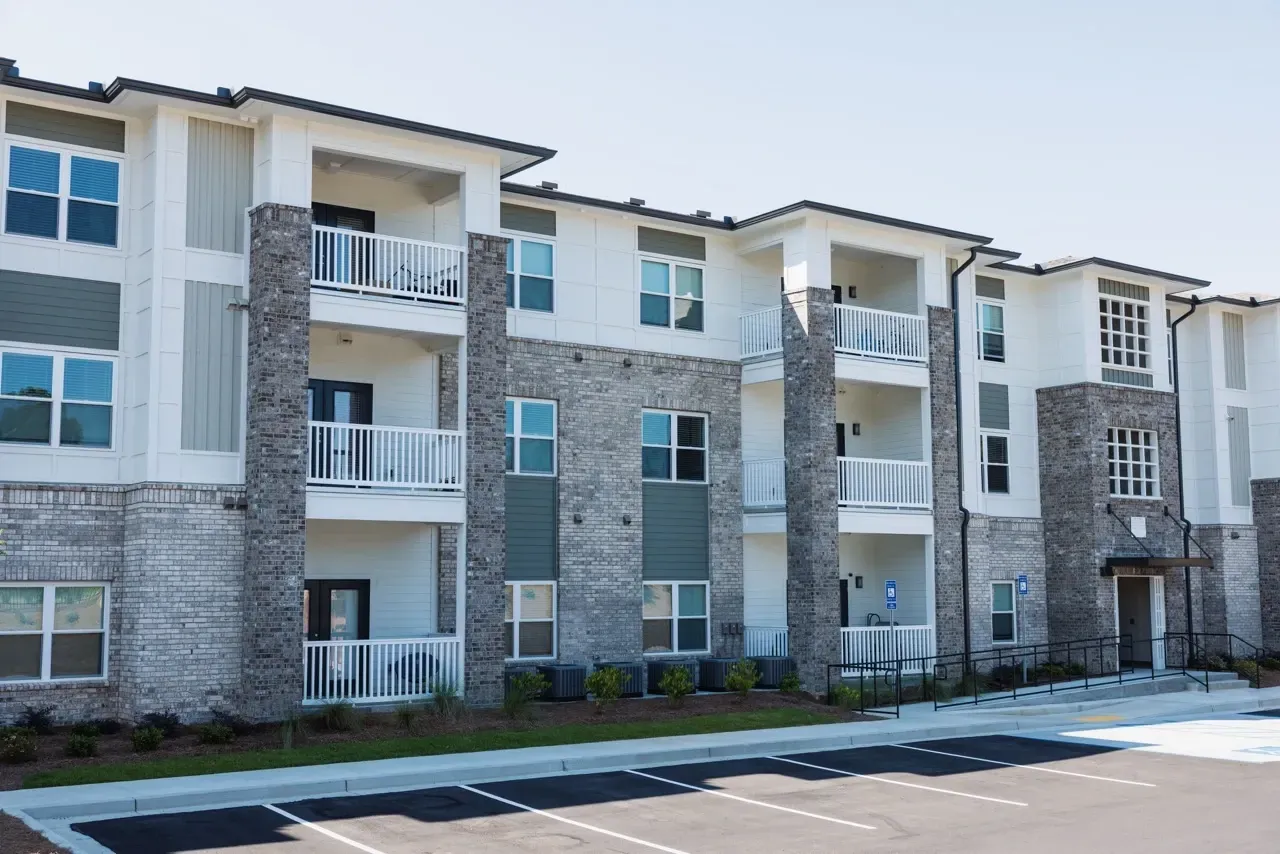 Exterior view of a modern, multi-story apartment building with brick columns, balconies, and a front parking lot.