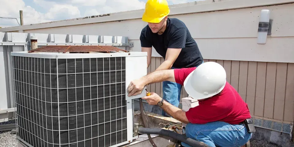 Two men are working on an air conditioner on the roof of a building.