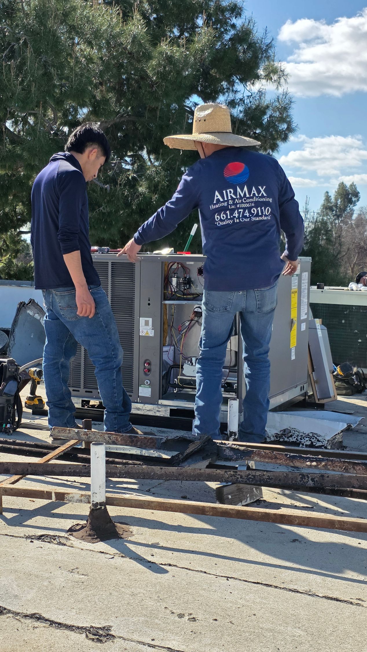 Electrician in gray shirt and red overalls smiling beside a white electrical panel, using a screwdriver.