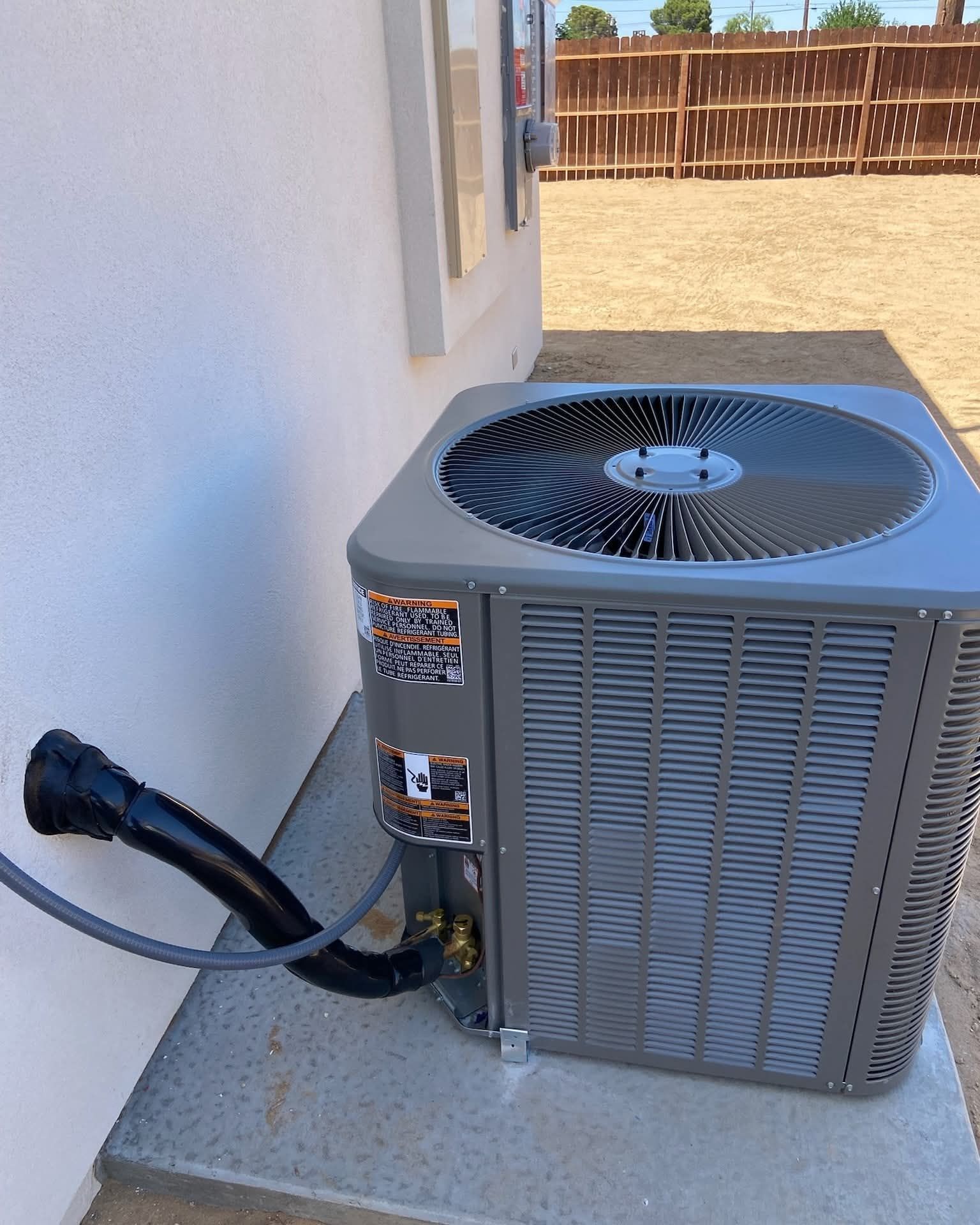 Outdoor air conditioner unit beside a white house wall with a hose connected, in a sunny yard.