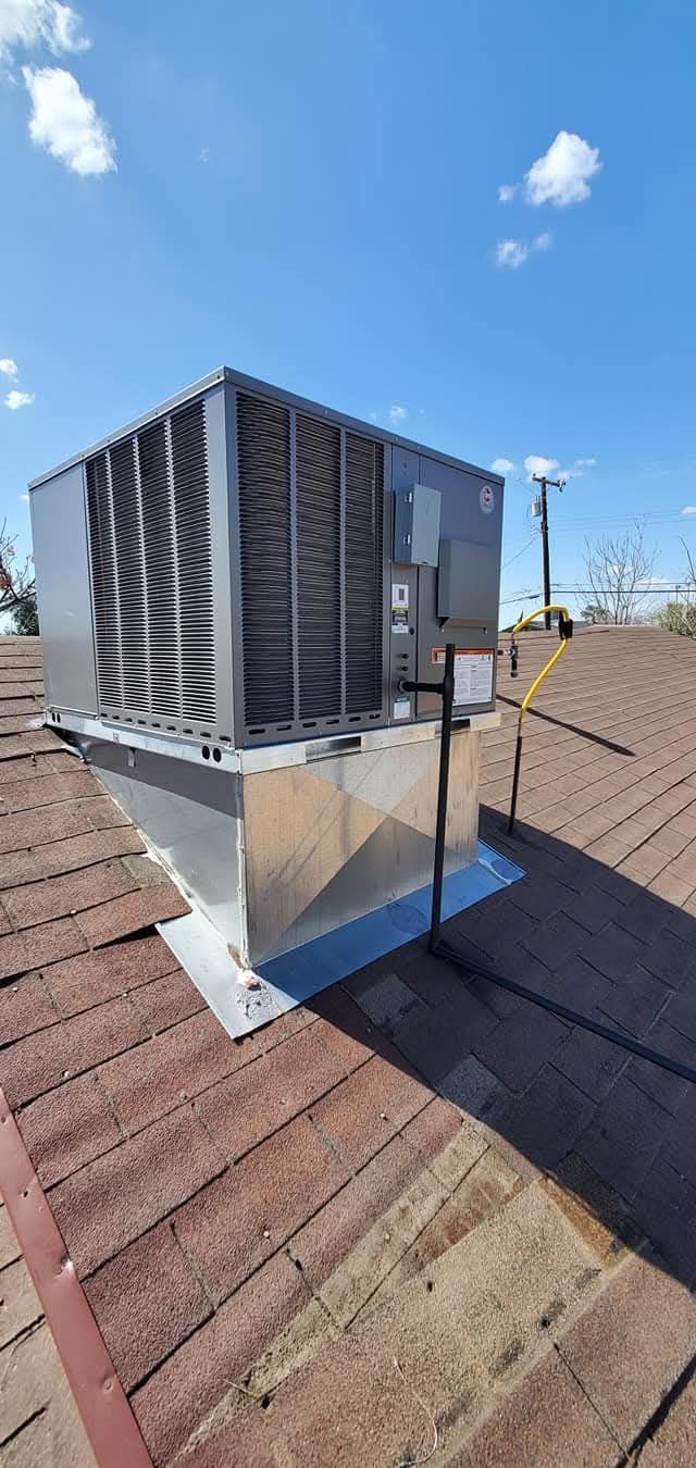 Rooftop HVAC unit with silver ductwork on a brown shingled roof under a blue sky.