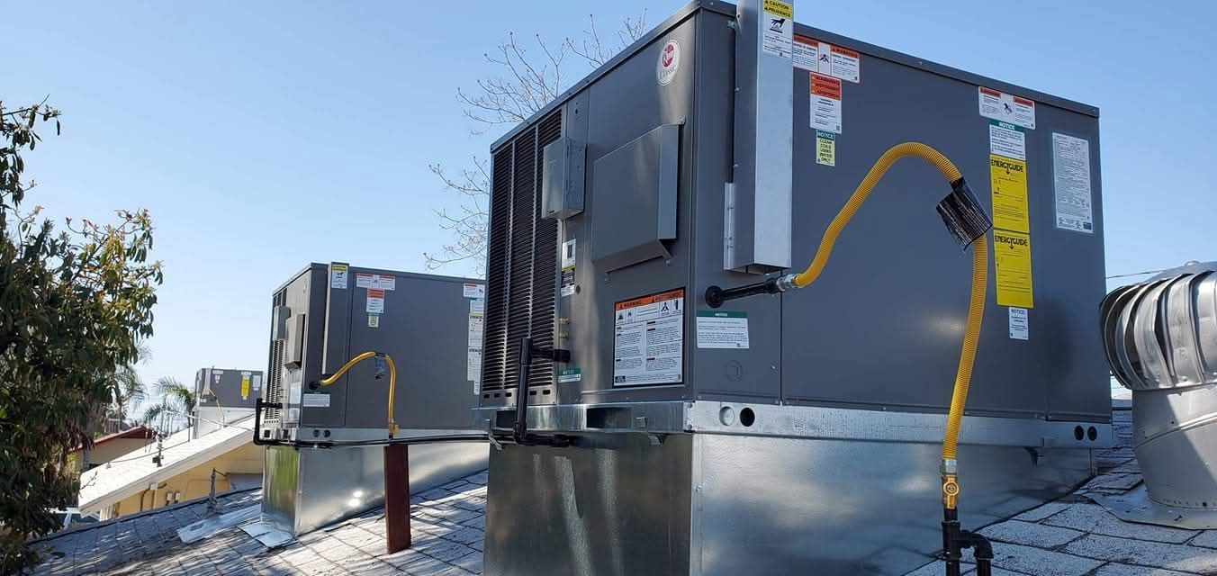 Two gray industrial HVAC units on a snowy rooftop, with yellow electrical conduit and vents nearby.