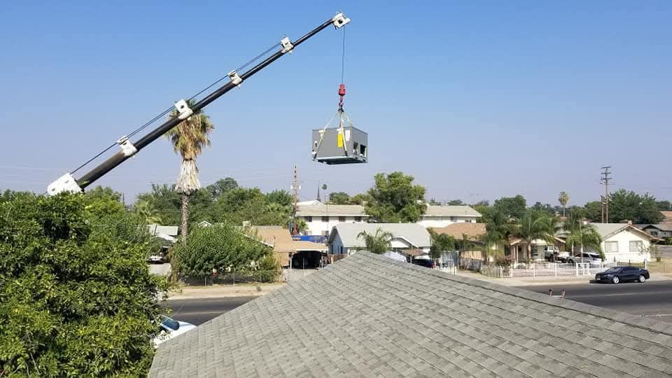 Crane lifting a small house over a suburban neighborhood under a clear blue sky