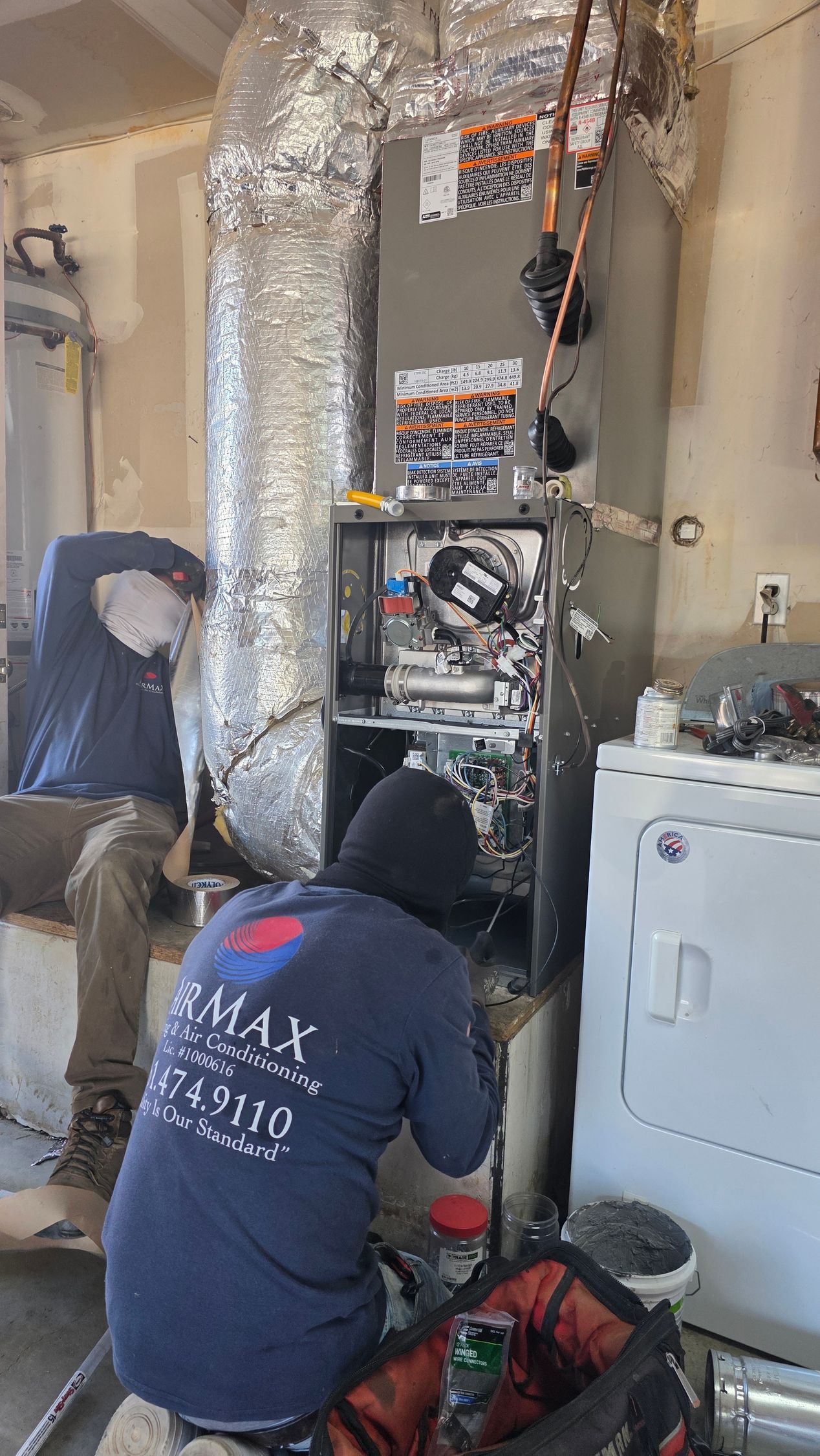 Technician adjusting the open furnace in a utility room, with ductwork and metal piping visible.