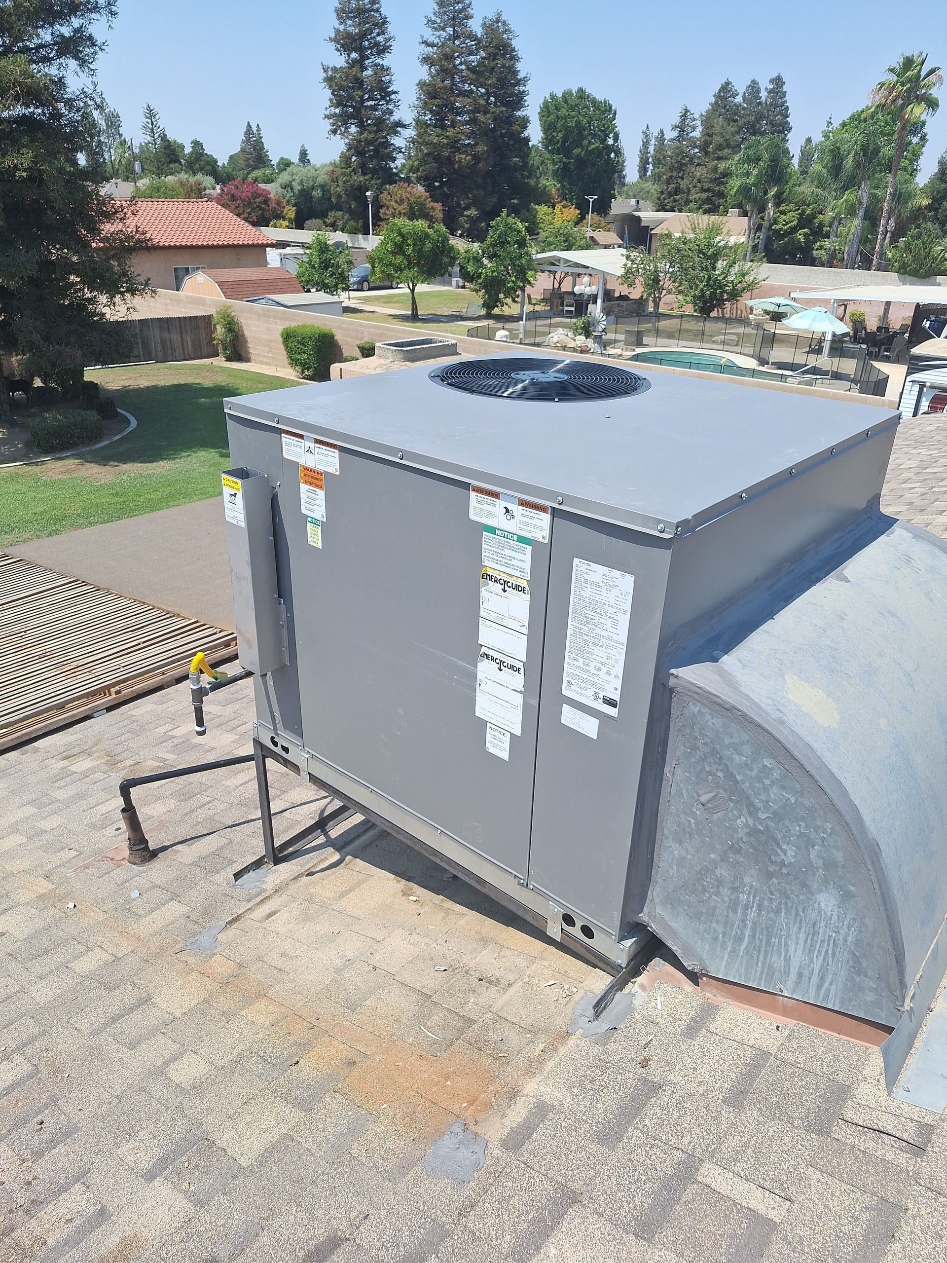 Large gray HVAC unit beside a curved metal duct on a rooftop with trees and houses in the background