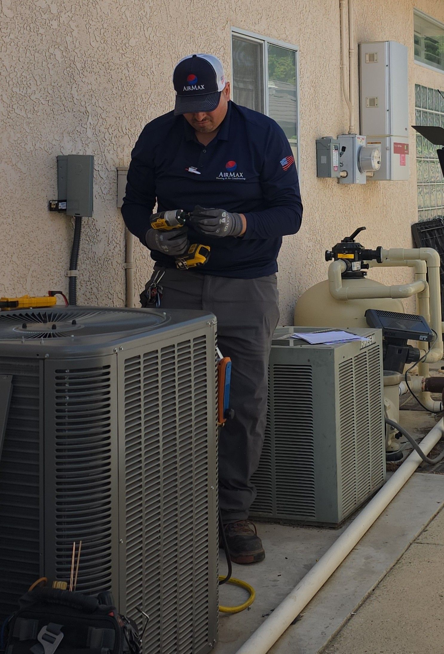 Outdoor HVAC unit beside a brick house, with black grille and red accent in a garden.
