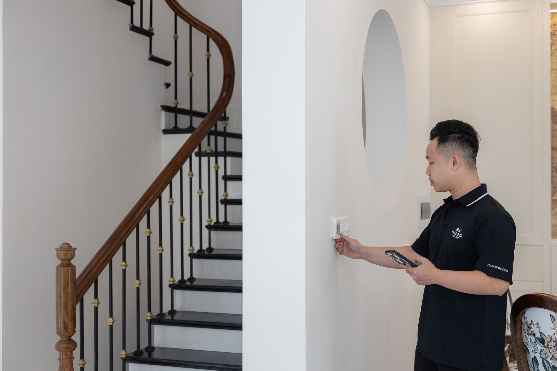 Man in a black shirt using a wall-mounted control panel beside a curved staircase inside a bright home