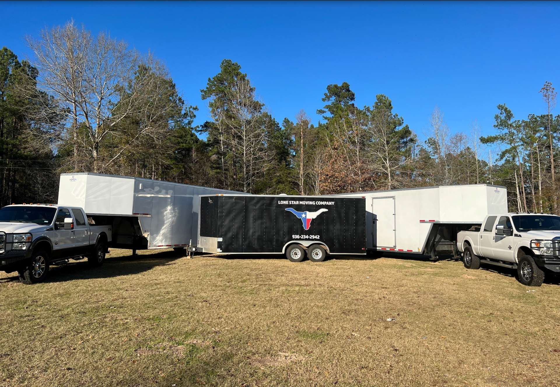 Several white and black trailers parked on a grassy field with trucks on each side under a clear blue sky