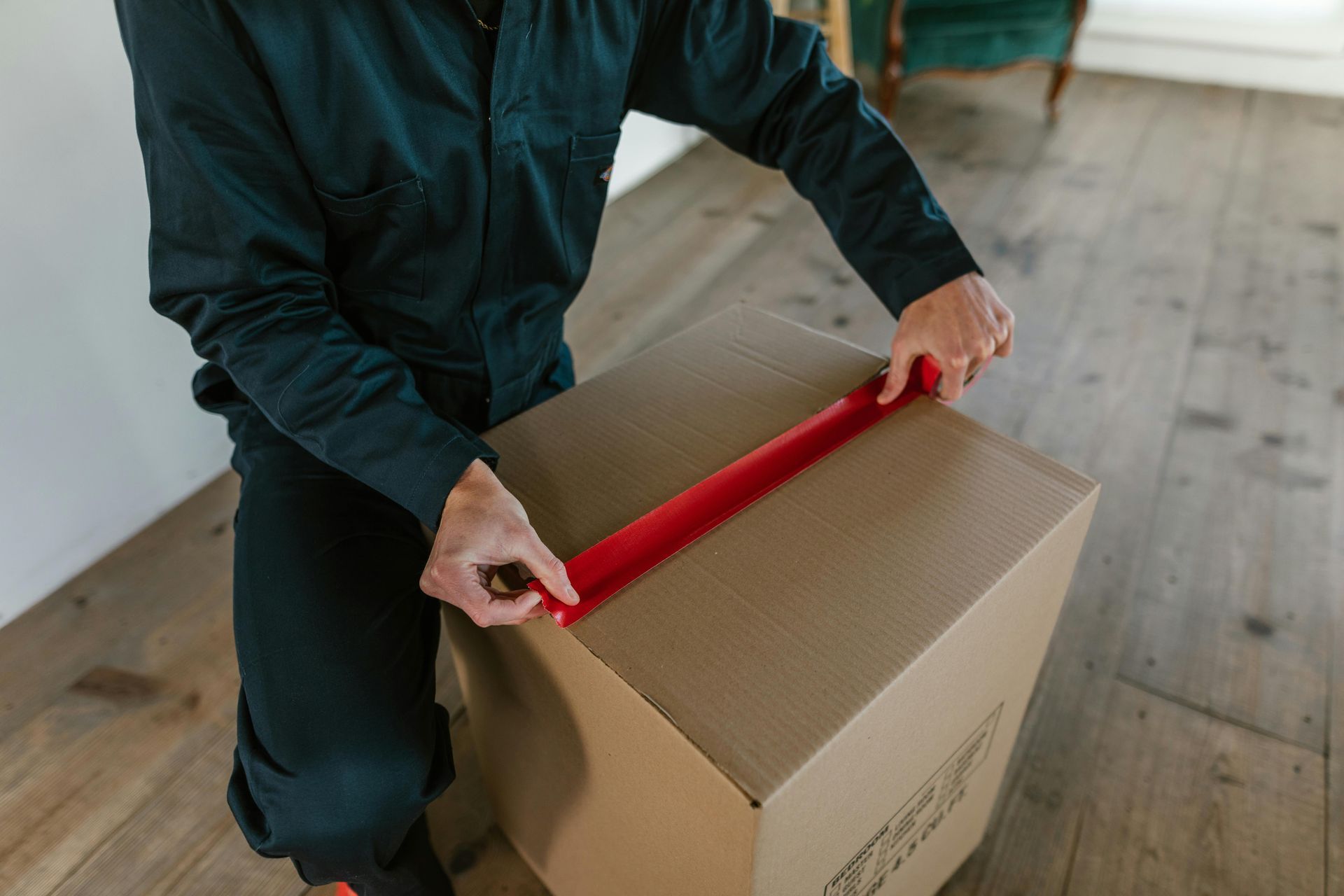 Person in dark clothing sealing a cardboard box with red tape on a wooden floor