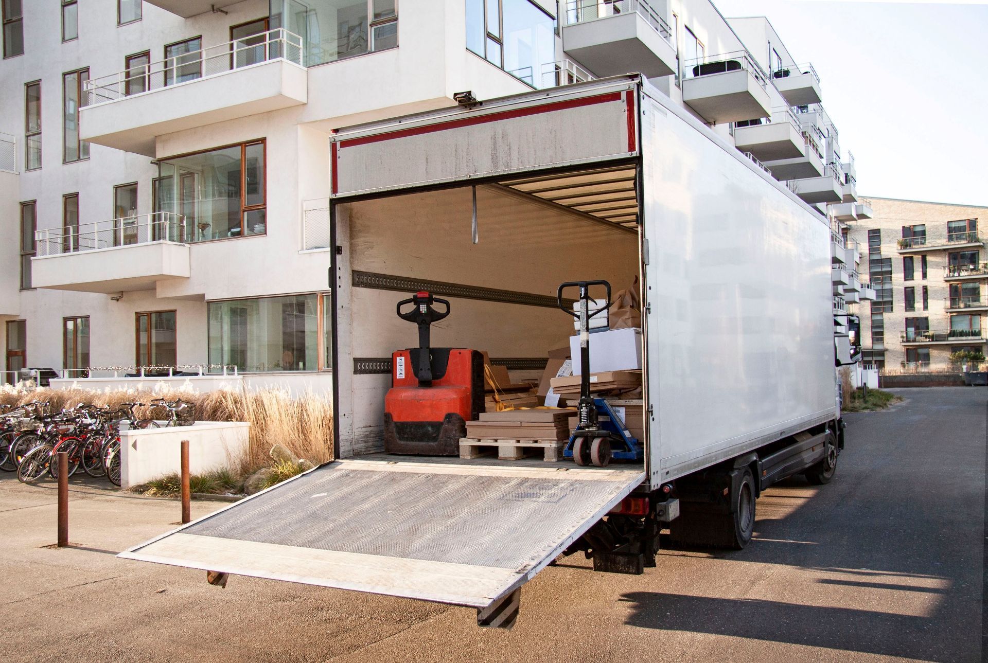 Moving truck with open ramp parked beside apartment buildings, carrying a red forklift inside.