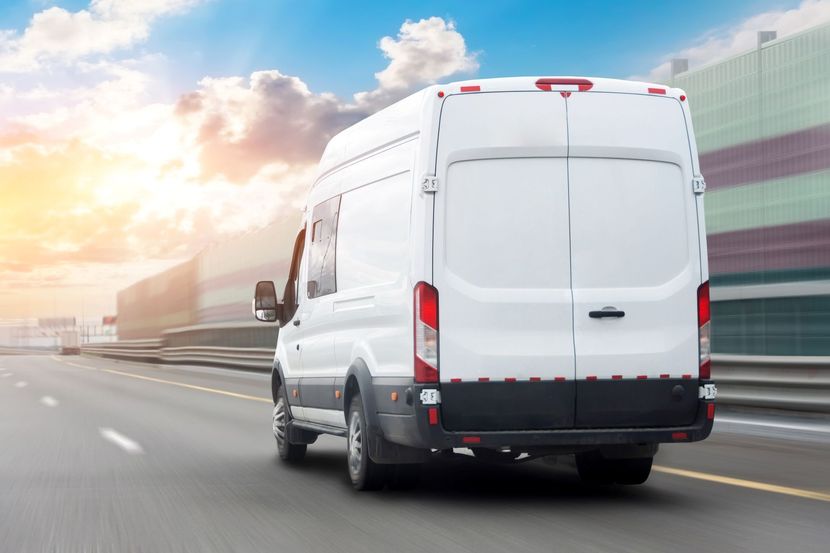 White delivery van driving on a highway beside a modern industrial building at sunrise