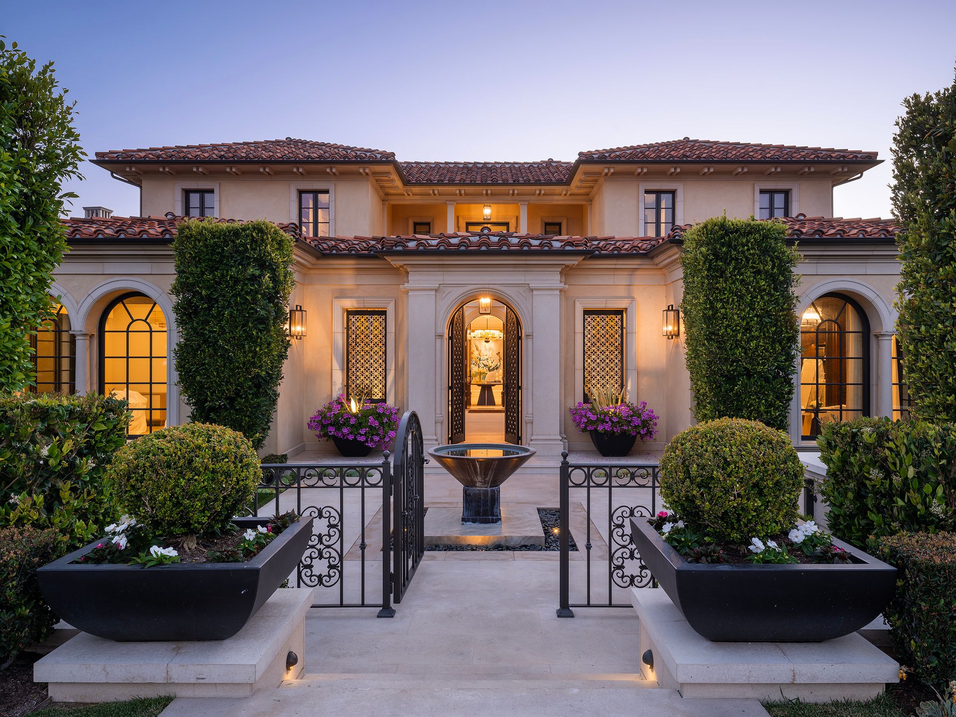 Elegant beige mansion with terracotta roof, gated entrance, and fountain, framed by manicured hedges and plants.