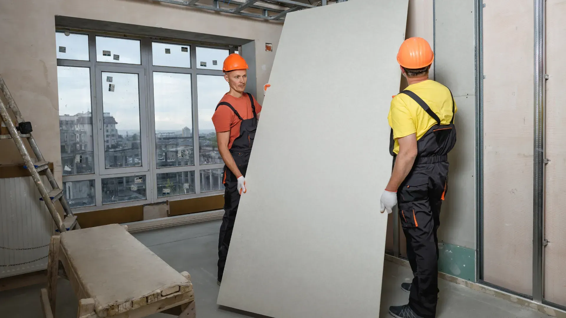 Two construction workers are carrying a large piece of drywall in a room.