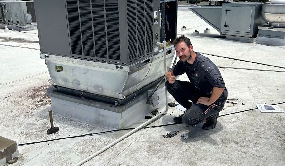 Technician kneeling by a rooftop HVAC unit, inspecting wiring on a snowy roof.