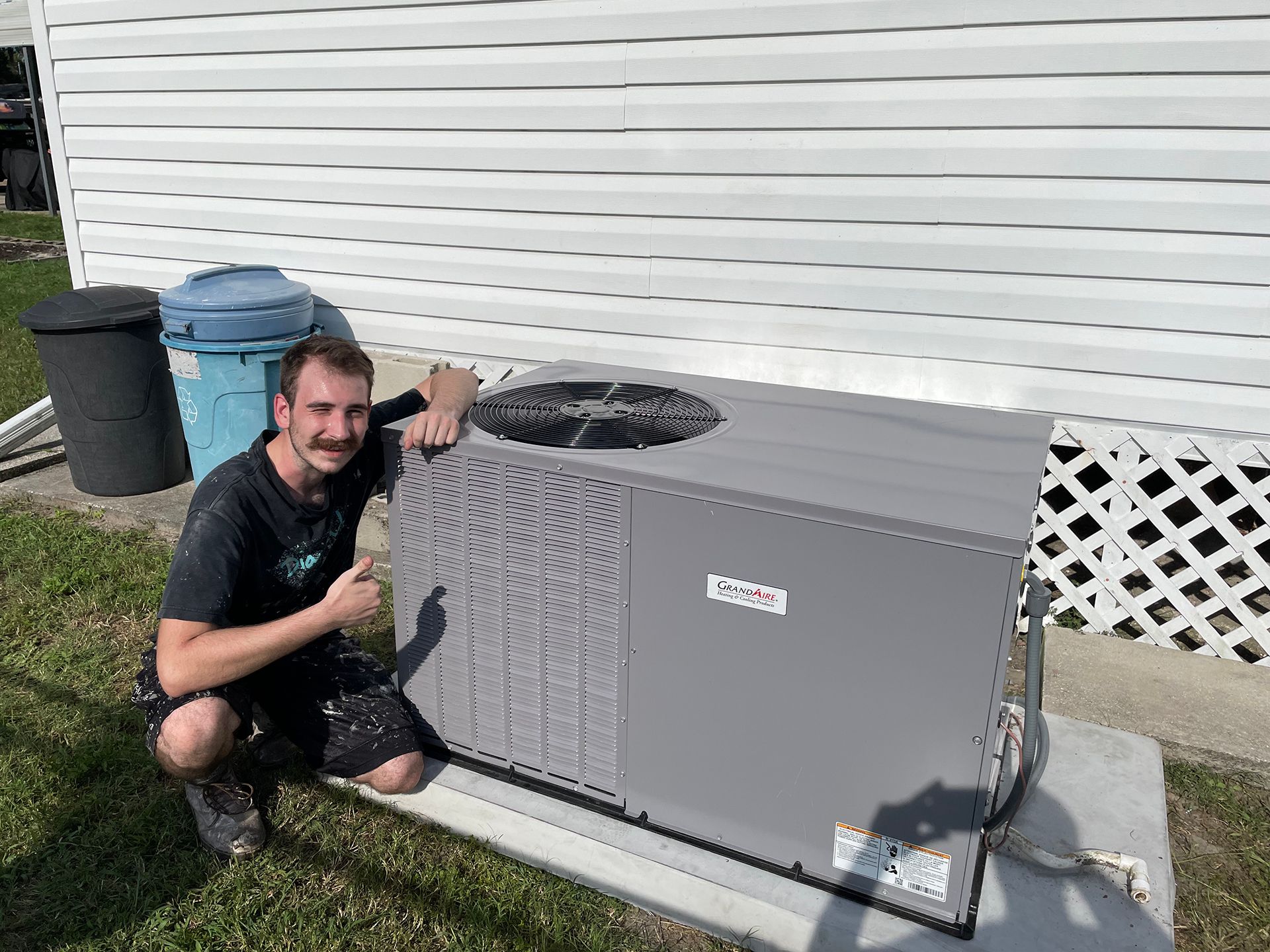 Man kneels beside a gray outdoor air conditioner unit next to a white house.