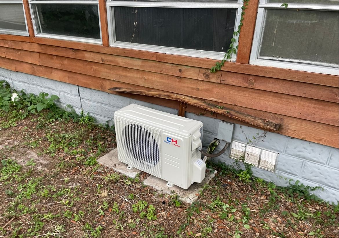 White outdoor HVAC unit beside a wooden house wall with windows and a concrete foundation