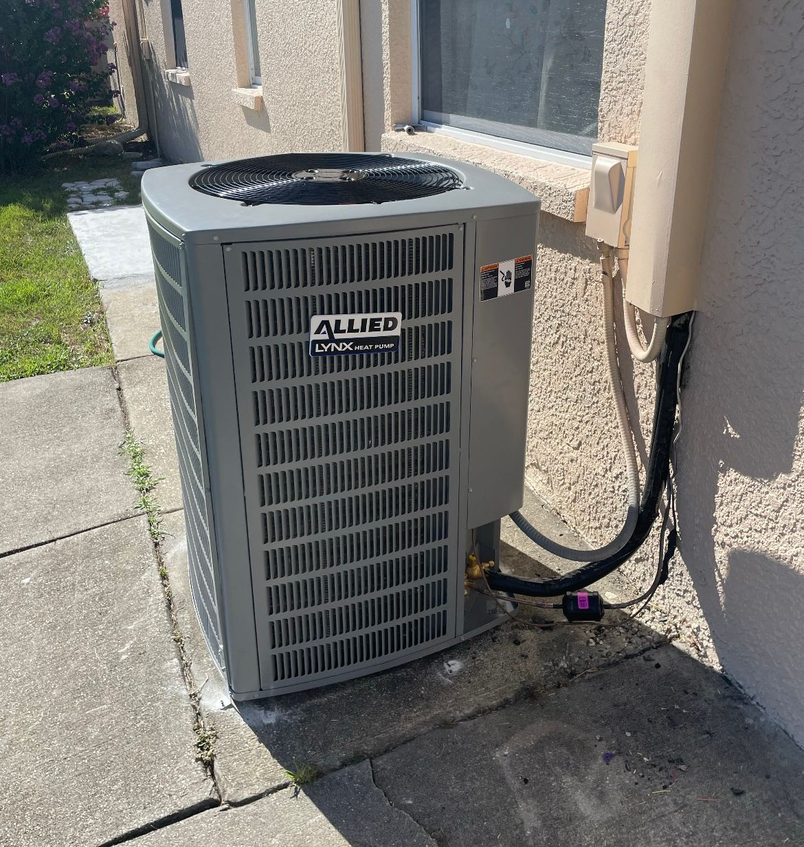 Outdoor air conditioner unit beside a stucco house wall, with a black hose on a concrete patio.