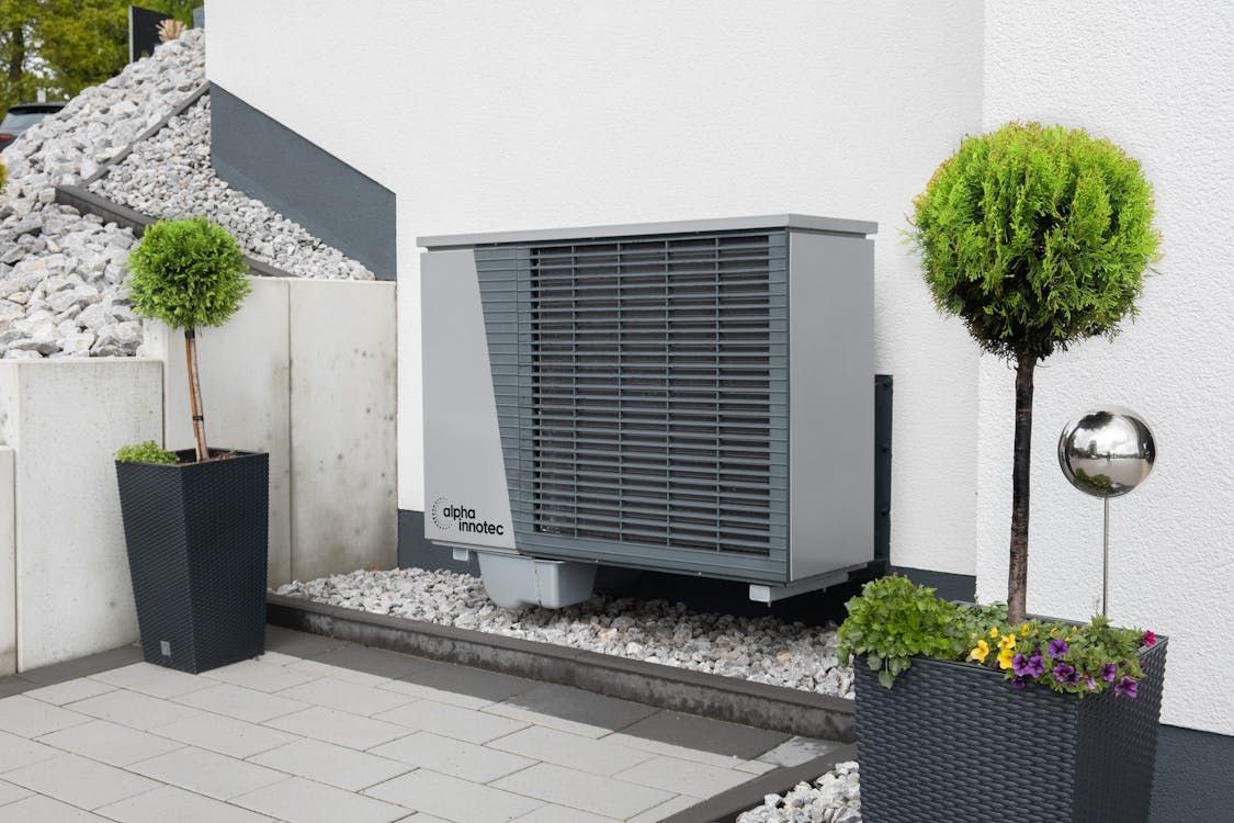 Outdoor heat pump unit beside white house wall, framed by black planters and a gravel bed.