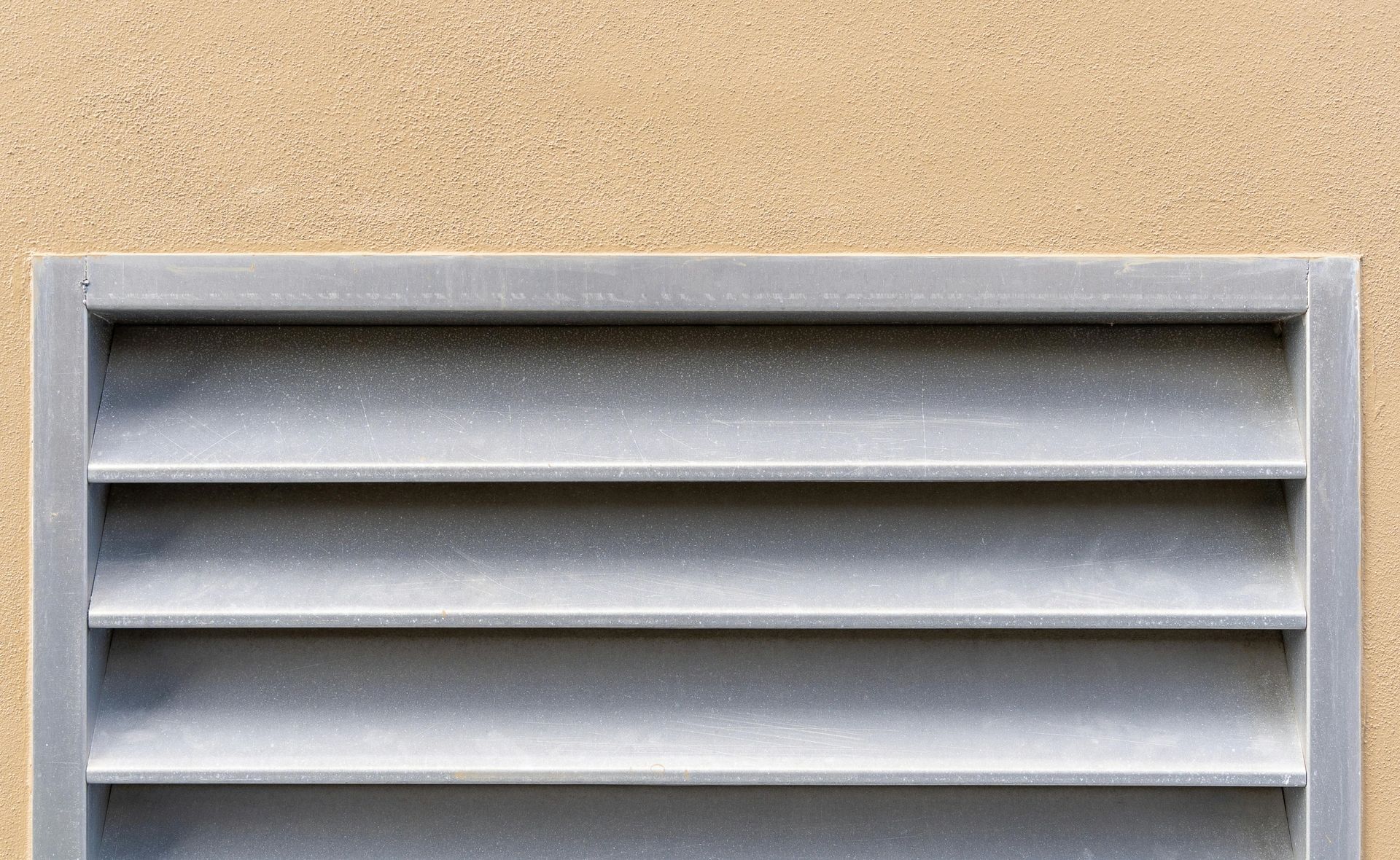 Gray metal vent with horizontal slats against a beige textured wall.