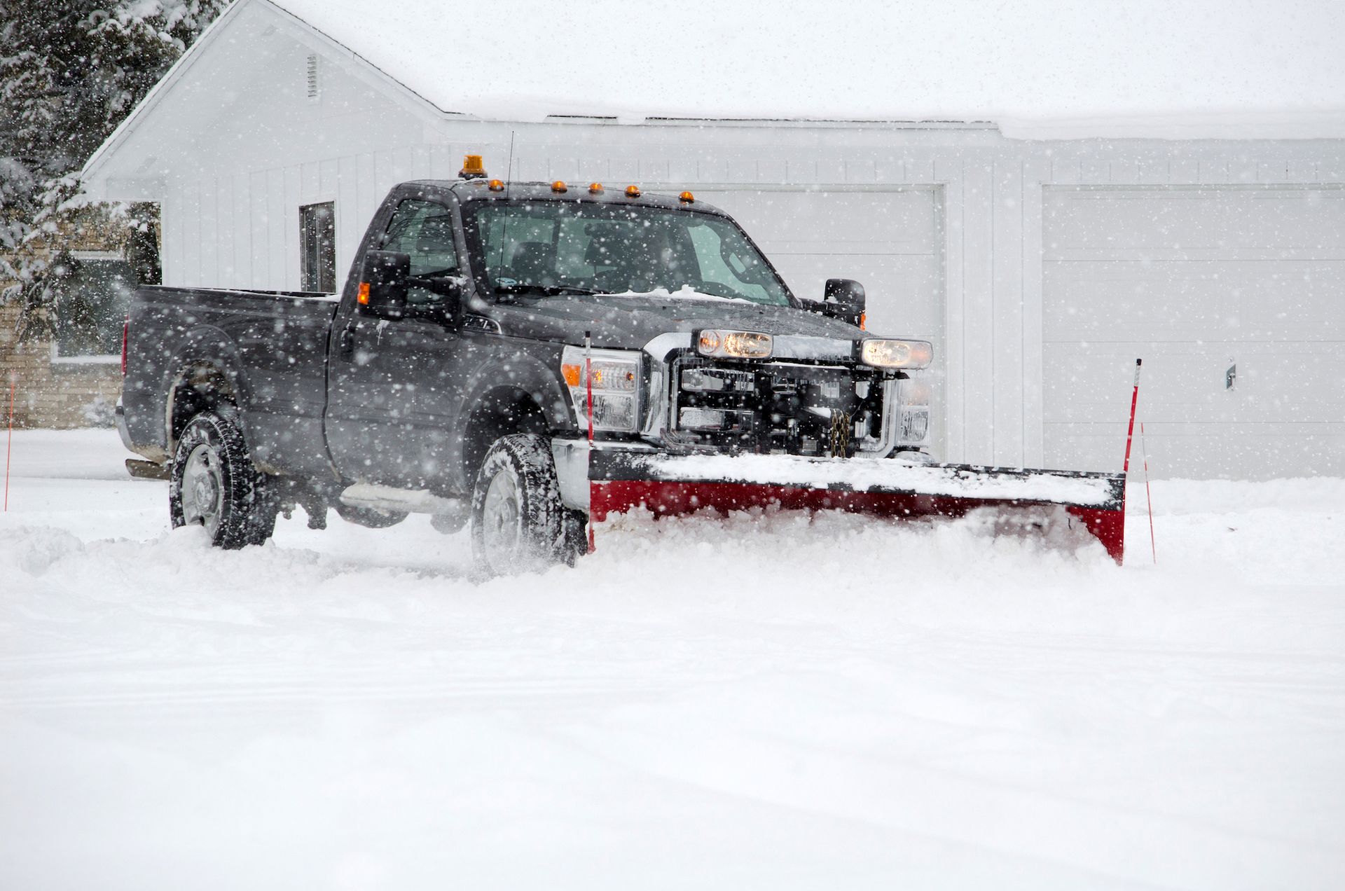 A dark-colored pickup truck with a large plow attachment clears deep snow in front of a white building.