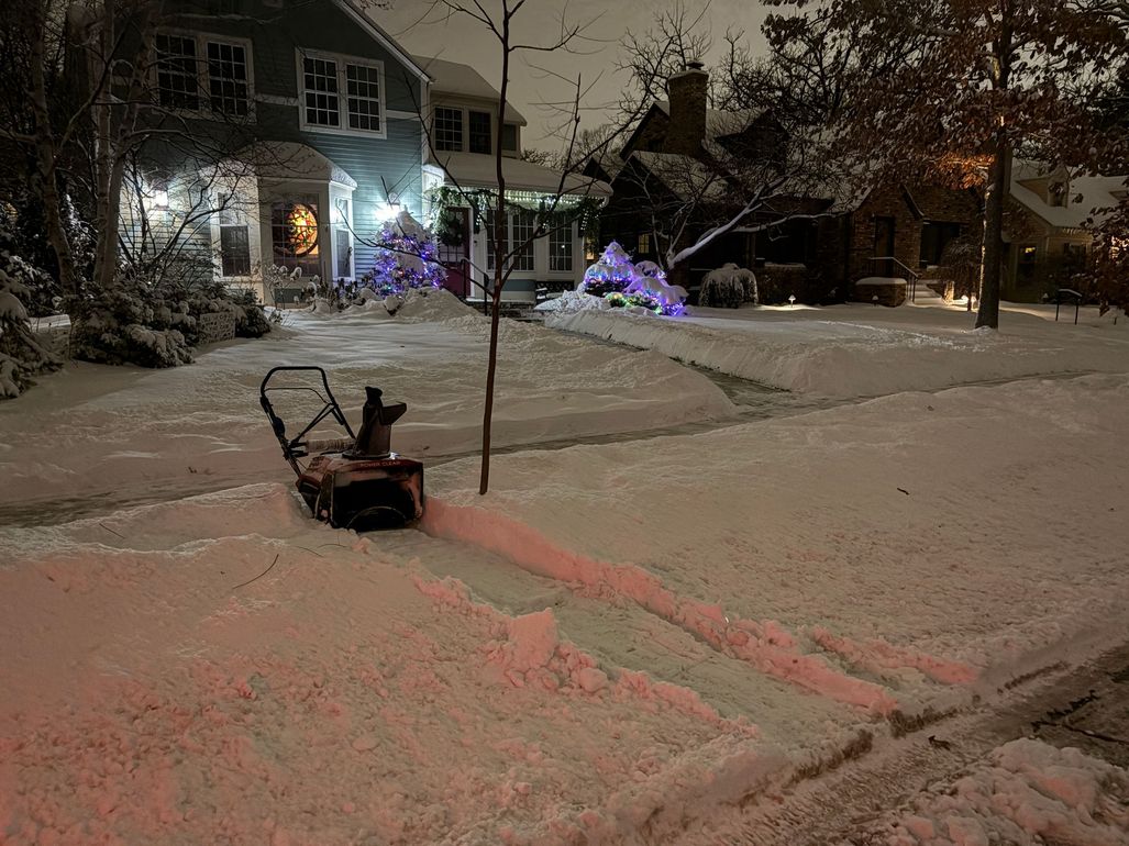 Snowy front yard at night with a snowblower clearing a path, house lit in the background.