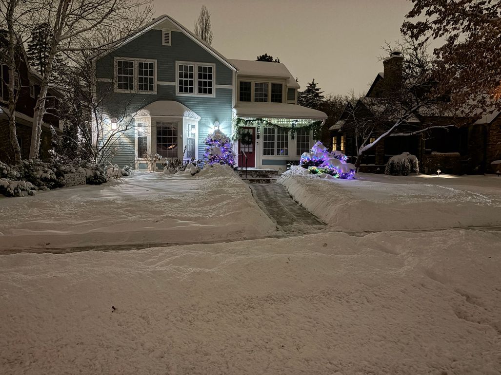Snow-covered house at night with glowing blue and purple holiday lights along the front walkway.